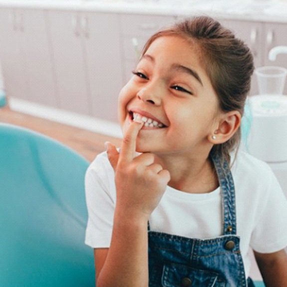 little girl sitting in dental chair and pointing to her teeth 